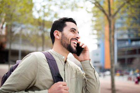 Side portrait of happy young north african guy walking outside with bag and talking on mobile phoneの写真素材