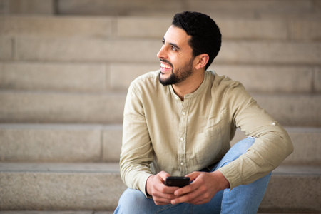Portrait of cheerful young arabic guy sitting on steps with cell phone looking away and smilingの写真素材