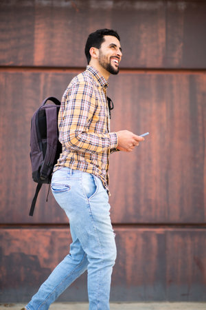 Side portrait of cheerful young arabic guy walking outside with bag and mobile phoneの写真素材