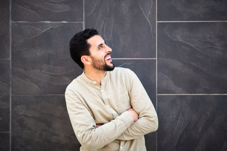 Portrait of young arabic man with arms crossed standing against black wall looking away and laughingの写真素材