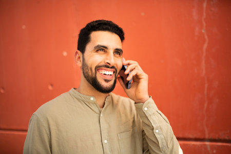 Close-up portrait of handsome young arabic man talking on phone against orange wallの写真素材