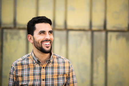 Portrait of handsome young arabic man standing against yellow wall looking away and smilingの写真素材