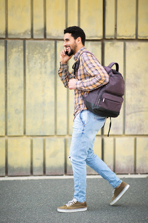 Full length side portrait of handsome young arab guy walking outside with bag and talking on mobile phoneの写真素材