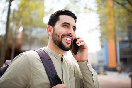 Portrait of handsome young middle eastern man walking outside with bag and talking on cell phoneの写真素材