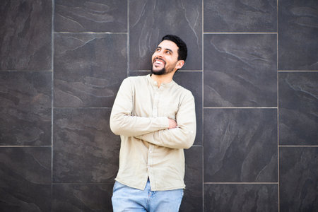 Portrait of confident young North African man standing with arms crossed against black tiled wall looking away and smilingの写真素材