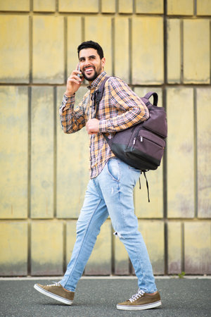 Full length portrait of cool young arabic man walking with bag and talking on phoneの写真素材
