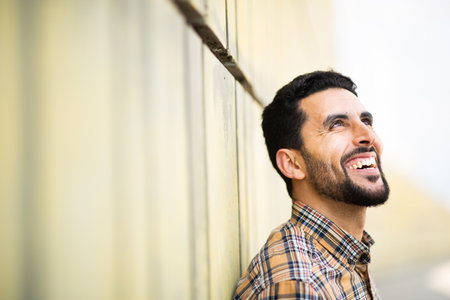 Close up side portrait of handsome young arab man looking away and smiling while leaning to a wall