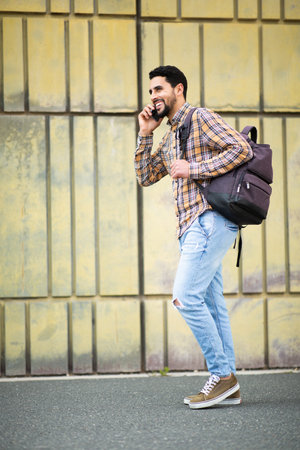 Full body side portrait of happy young arabic guy walking with bag and talking on cell phoneの写真素材
