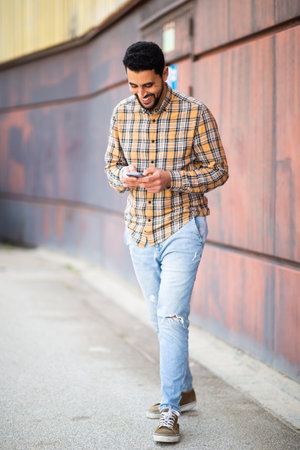 Full length portrait of young middle eastern man walking outside and using mobile phoneの写真素材