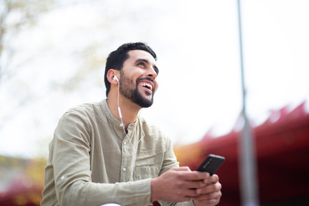 Portrait of smiling man sitting outside with earphones enjoying listening to music from his cell phoneの写真素材