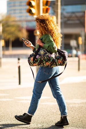 Full length side portrait of young travelling woman walking outside with bag reading text message on...