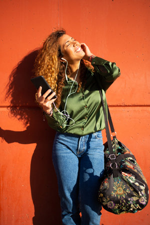 Portrait of young latin woman listening music on smart phone leaning to orange wallの写真素材