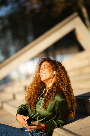 Portrait of beautiful young woman enjoying listening music from mobile phone sitting outside on stepsの写真素材