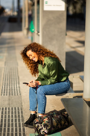 Side portrait of cheerful young woman texting on cellphone while waiting for public transport in the cityの写真素材