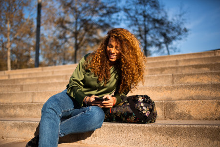 Portrait of young hispanic woman sitting on steps outside texting on mobile phoneの写真素材