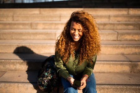 Portrait of travel woman with bag and cellphone sitting on steps outside and smilingの写真素材