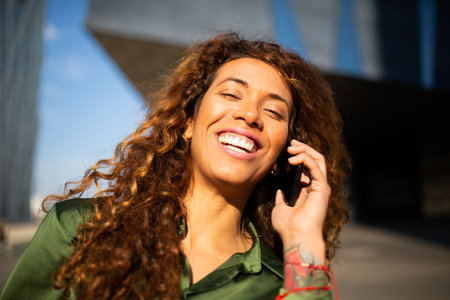 Close up portrait of cheerful young latin woman talking on cellphone outside in the cityの写真素材