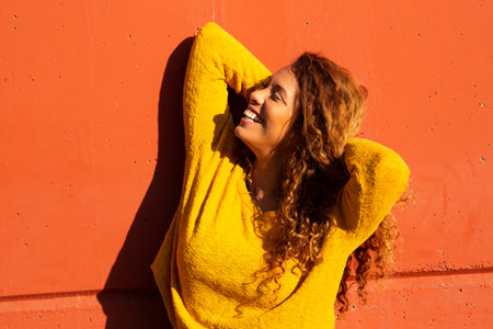 Portrait of young hispanic woman leaning against orange wall looking away with hands behind head and smilingの写真素材
