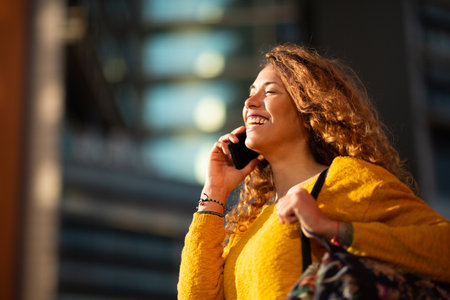 Portrait of cheerful young latin woman with bag talking on mobile phone while walking outside in the cityの写真素材