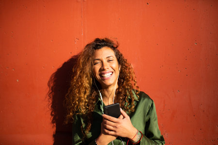 Portrait of laughing young latin woman with cell phone and earphones standing against orange wallの写真素材