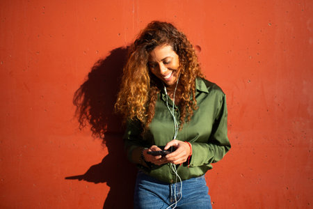 Portrait of smiling young woman listening music with earphones and smart phone leaning to orange wallの写真素材