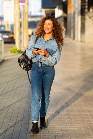 Full body portrait of smiling young latin woman walking with bag and mobile phone outside in the cityの写真素材