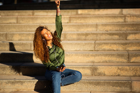 Portrait of happy hispanic woman sitting on steps outside enjoying listening to musicの写真素材
