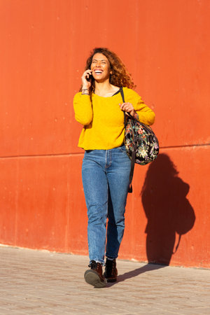 Full length portrait of young woman walking with bag and talking on mobile phone outside on a sidewalkの写真素材