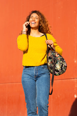 Portrait of cheerful latin woman walking with bag and talking on mobile phone against orange wallの写真素材