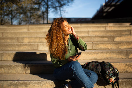 Portrait of beautiful latin woman sitting on steps outside listening to music with earphones and phoneの写真素材