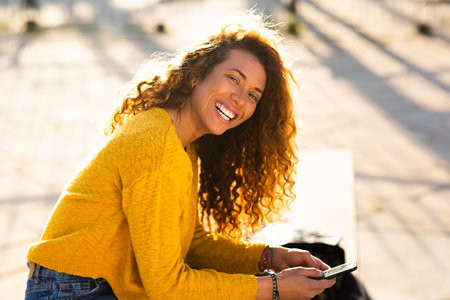Portrait of young latin woman with cellphone sitting outside in the cityの写真素材