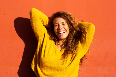 Portrait of beautiful young hispanic woman in yellow top posing against orange wall with hands behind headの写真素材