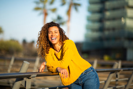 Portrait of smiling young hispanic woman leaning to a railing looking at camera and laughing outside in the cityの写真素材