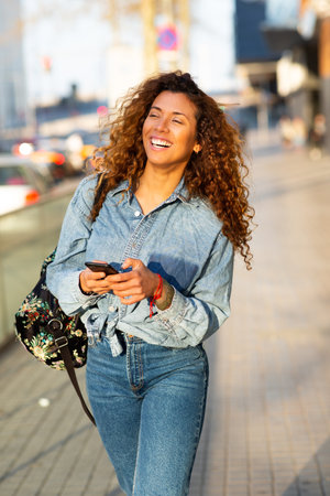 Portrait of beautiful young latin female commuter walking with bag and mobile phone outside in the cityの写真素材