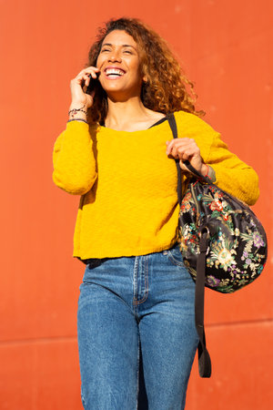 Portrait of young latin woman talking on mobile phone while walking against orange wallの写真素材