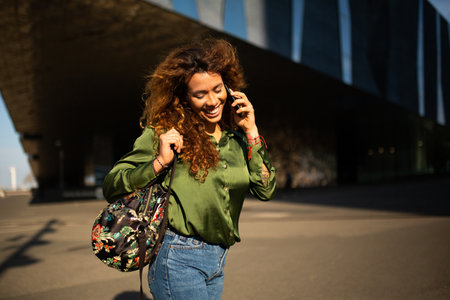 Portrait of cheerful hispanic woman talking with mobile phone outdoors in cityの写真素材