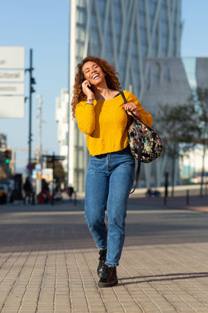 Full body portrait of happy young latin woman walking and talking on mobile phone outside in the cityの写真素材