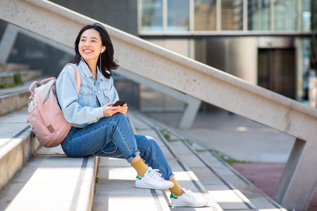 Portrait smiling young asian woman sitting with mobile phone outsideの写真素材