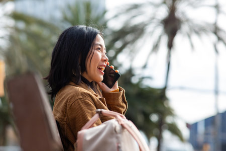 Side portrait smiling young asian woman talking with cellphone while sitting on bench in cityの写真素材