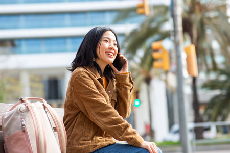 Portrait smiling young asian woman talking with mobile phone while sitting on benchの写真素材