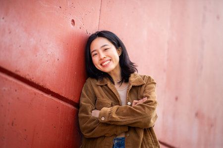 Portrait happy young asian woman with arms crossed leaning against wallの写真素材