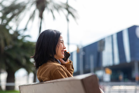 Side portrait asian woman talking with cellphone while sitting on bench in cityの写真素材