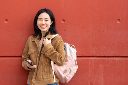 Portrait smiling young woman standing with mobile phone and bag by wallの写真素材