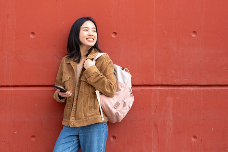 Portrait young woman standing with mobile phone and bag by wallの写真素材