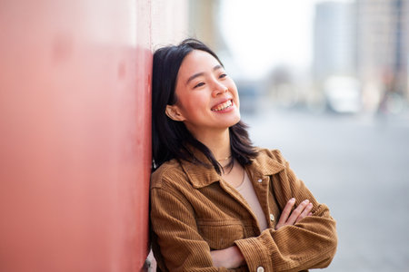 Portrait smiling young asian woman with arms crossed leaning against wallの写真素材