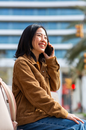 Side portrait smiling young asian woman talking with mobile phone while sitting on benchの写真素材