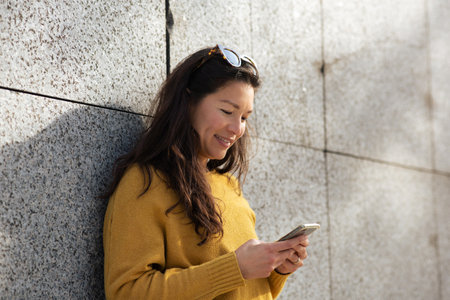 Portrait of happy asian woman leaning against wall and using mobile phone outsideの写真素材
