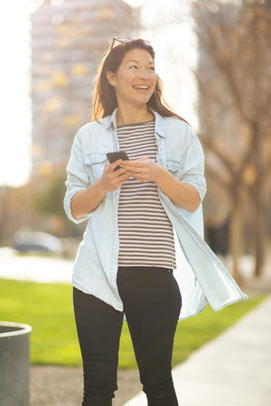Portrait of smiling asian woman with cell phone and looking away walking outside in the city