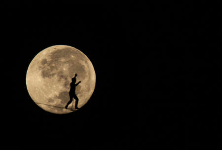 silhouette of a man walking a tightrope in landscape and in the background the full moonの写真素材