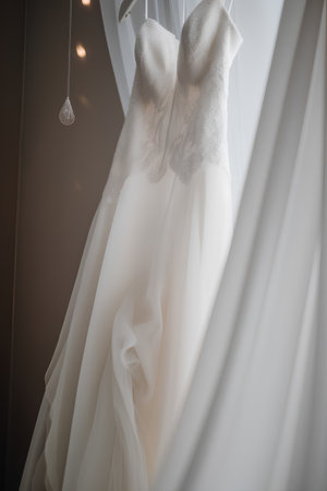 Wedding dress hanging on a white curtain in a room.の素材